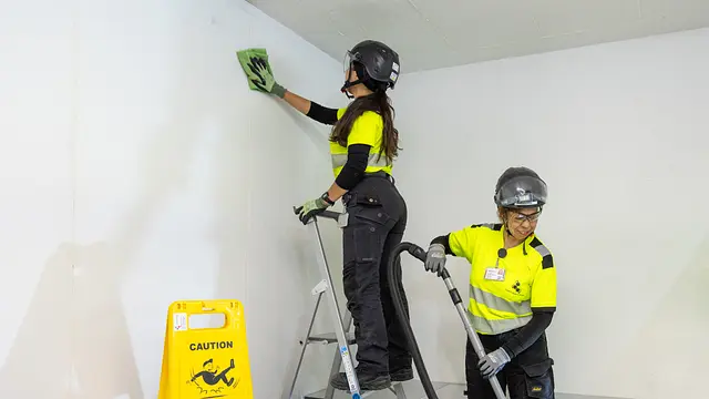 Two team members clean the walls of a data center, one on a ladder with a cloth, one with a vacuum, a warning sign on the raised floor in front.