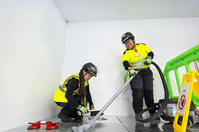 Two workers in helmets, safety glasses and high-visibility clothing clean a raised floor with a hand brush and a vacuum, with barriers and a warning sign at the edge.